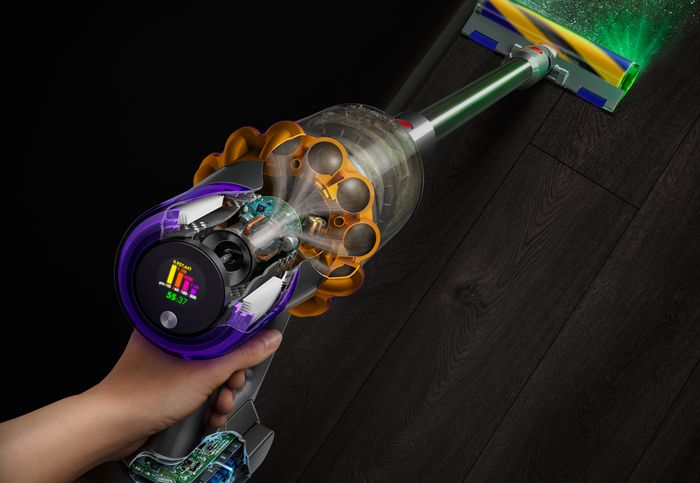 Ruka drží vysavač Dyson V15 Detect Absolute v řezu, hubice se zeleným světelným paprskem svítí na tmavou dřevěnou podlahu