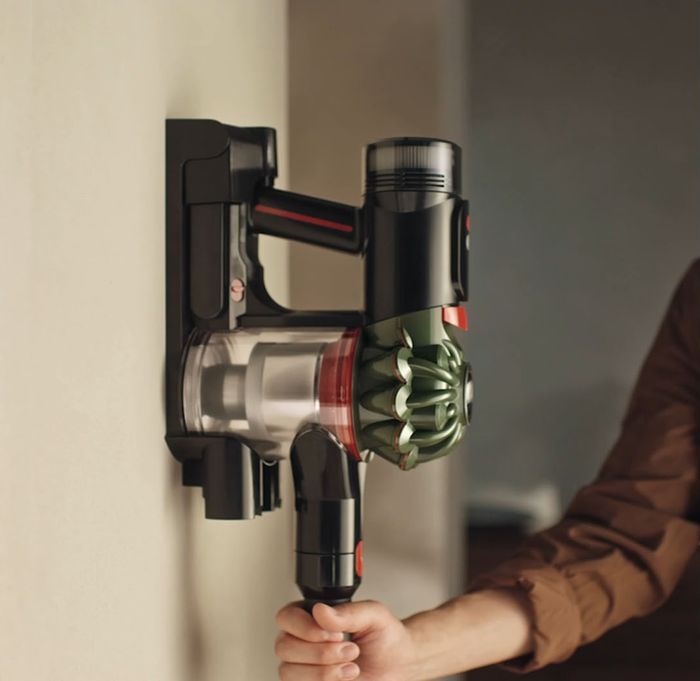 Vysavač Dyson V8 Cyclone 2026 zavěšený v nástěnném úchytu na světlé stěně, ruka drží jeho rukojeť