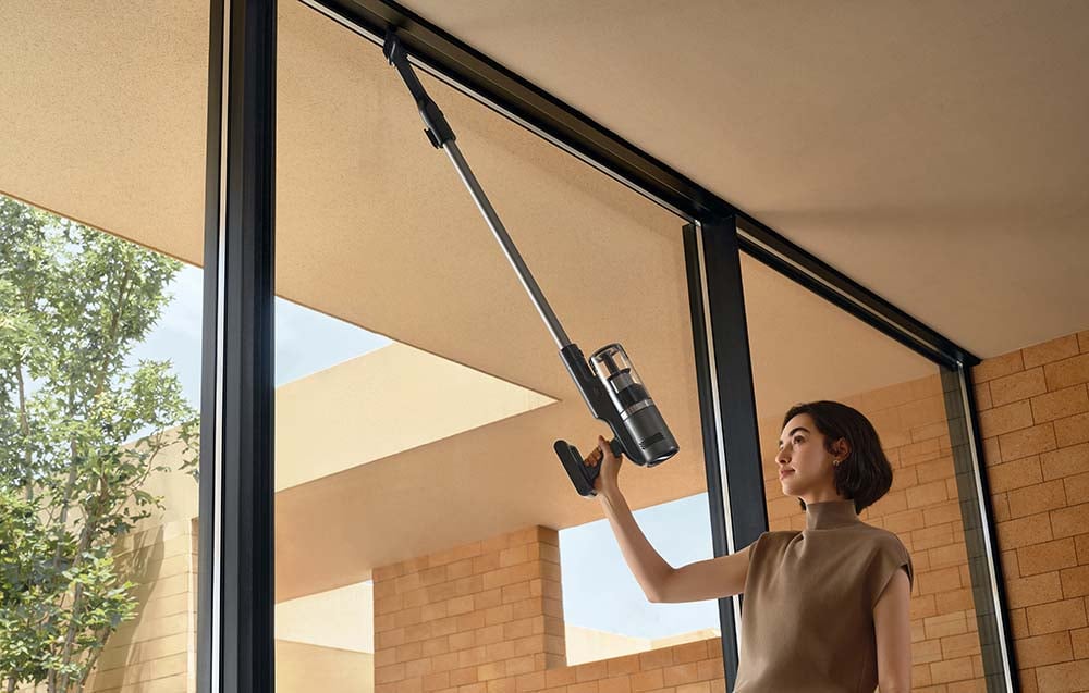 A woman vacuums the window frames with an upright vacuum cleaner.