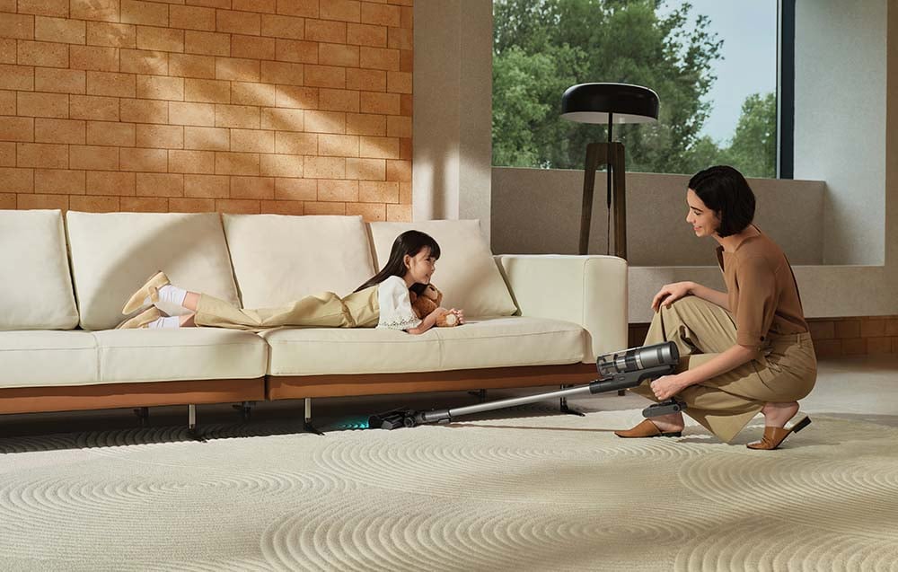 A woman is vacuuming the carpet under the couch, a girl is lying on the couch, holding a teddy bear in her hand.