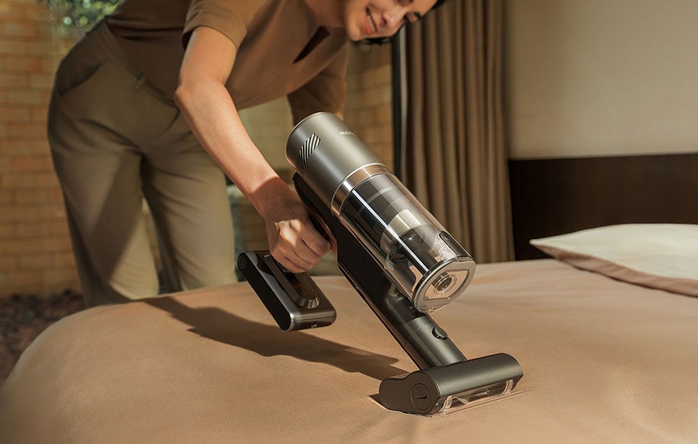 A smiling woman vacuums the bed with a handheld vacuum cleaner.