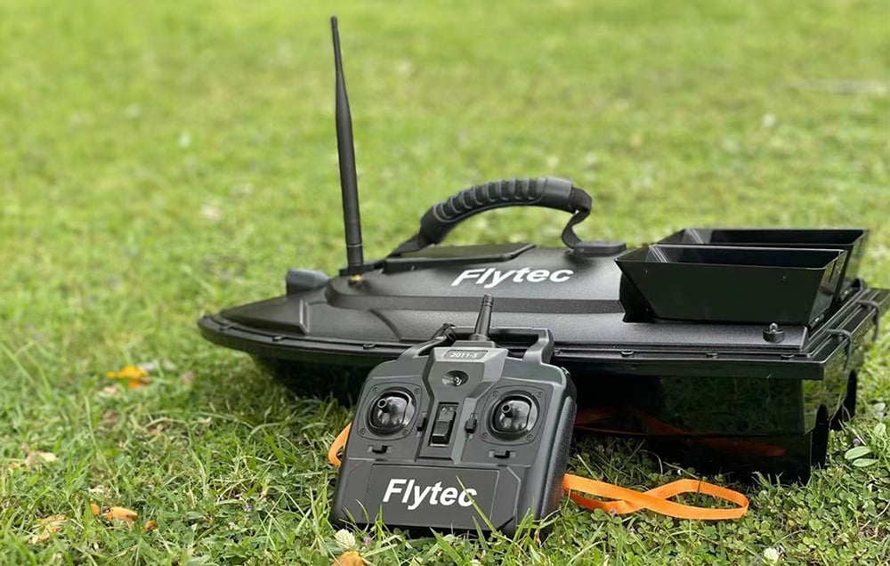 Černá loďka s návnadou Flytec s dálkovým ovládáním ležící na zelené trávě