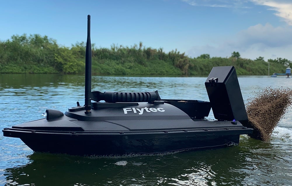 Člun na návnady Flytec s přihrádkou na návnady plovoucí na jezeře se zeleným břehem v pozadí