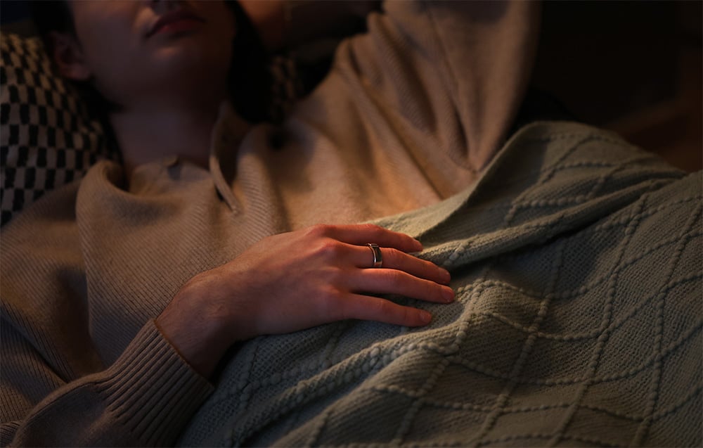 Osoba ležící pod přikrývkou s rukou položenou na břiše, na prstě má stříbrný prsten.