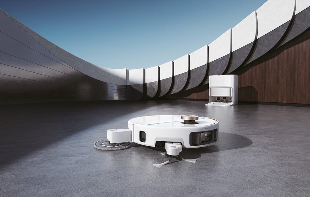 Bílý úklidový robot s vysunutým mopovacím modulem při práci na podlaze, v pozadí s dokovací stanicí MOVA