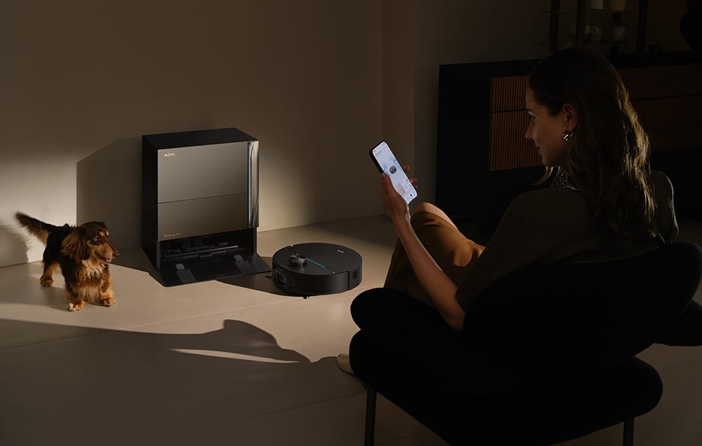 Žena sedí v křesle se smartphonem v ruce; vlevo je jezevčík, dokovací stanice a úklidový robot