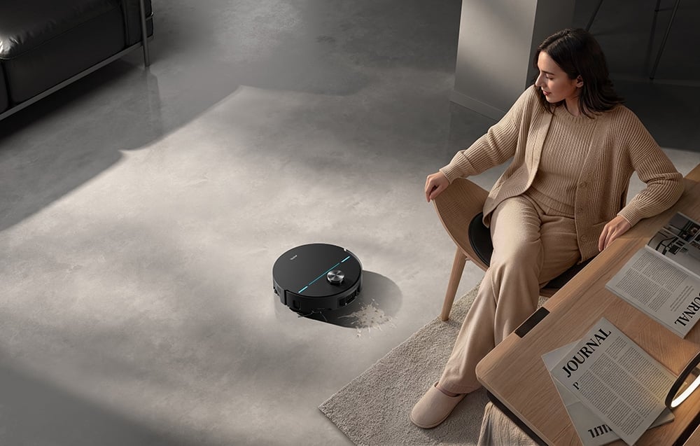 Žena sedí na židli u stolu; vedle ní úklidový robot myje podlahu