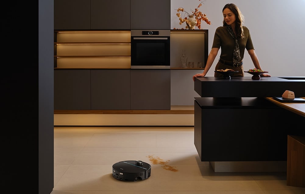 Žena stojí v kuchyni opřená o kuchyňský ostrůvek; vedle ní úklidový robot odstraňuje skvrnu z podlahy